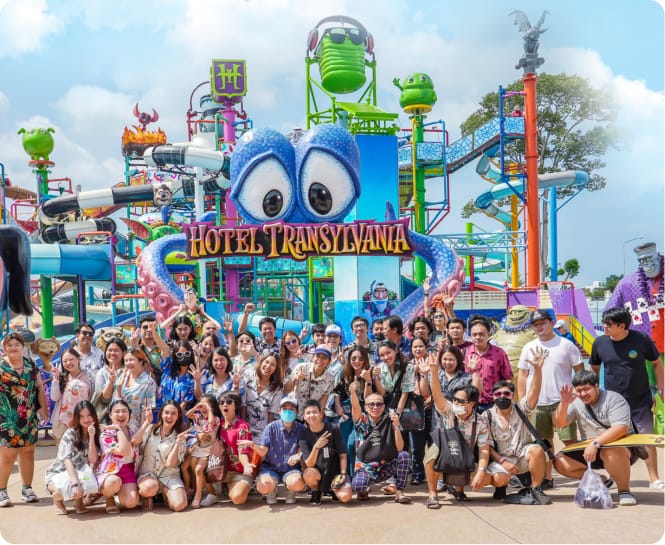 A group of people are posing in front of the Hotel Transylvania aqua park. Giant splash buckets and water slides can be seen in the background.