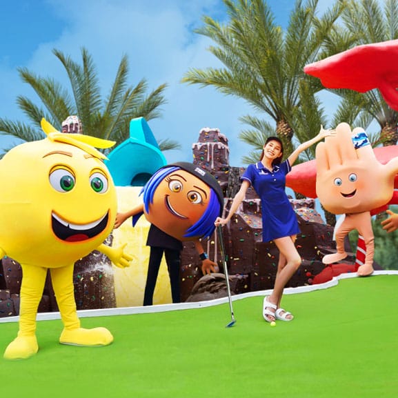 A woman is posing with the characters from The Emoji Movie on a mini golf's putting green.